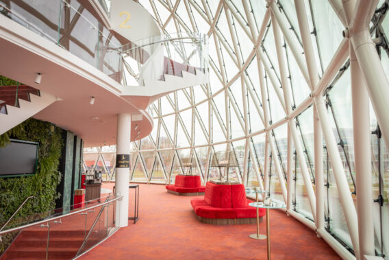 Foyer Begane Grond. Via de glazenkoepel kan je genieten van daglicht en het uitzicht naar buiten. Rode fluwelen banken bieden comfort tijdens het wachten.