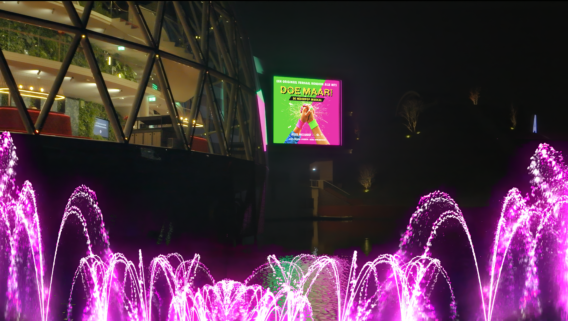 Buitenaanzicht van het AFAS Theater Leusden in avondsferen, met glinsterende roze verlicht fontein en Doe Maar artwork in de achtergrond.